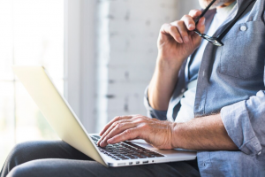 Uma pessoa sentada está usando um laptop, segurando óculos em uma mão e parecendo refletir ou fazer uma pausa da tela. A pessoa veste uma camisa jeans e calças escuras, e o cenário borrado ao fundo sugere um ambiente interno iluminado naturalmente